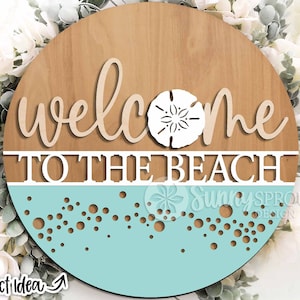 May include: A round wooden sign with the text "Welcome to the Beach" in white lettering. The sign has a light blue background with a white sand dollar and white dots.