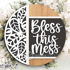 May include: A round wooden sign with a decorative white cutout design on the left and the words "Bless this Mess" in white on a dark gray background. The sign has a natural wood grain finish and is surrounded by white flowers.