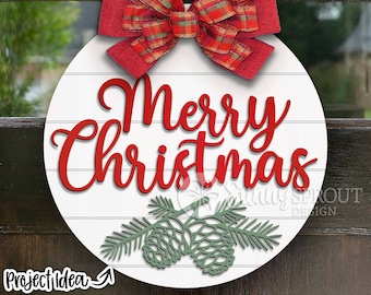 Pode incluir: Um sinal redondo de madeira branca com letras vermelhas que dizem "Feliz Natal". O sinal é decorado com um laço vermelho xadrez e pinhas verdes.