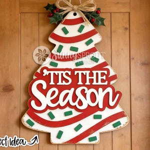May include: A white wooden Christmas tree door hanger featuring red diagonal stripes and small green rectangular accents. The text 'TIS THE SEASON' is displayed in large white letters on a red background. A jute bow and holly leaves top the piece.