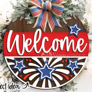 May include: A round wooden sign with a red, white, and blue patriotic theme. The sign says "Welcome" in white letters and features a firework design with stars.