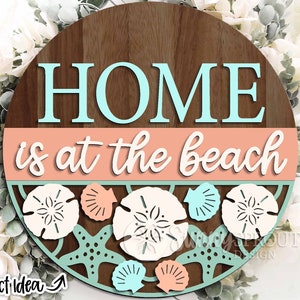 May include: A round wooden sign with the text "HOME is at the beach" in white and blue lettering. The sign is decorated with seashells, sand dollars, and a starfish in shades of blue, white, and orange.