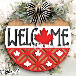 May include: A round wooden welcome sign with a Canadian theme. The sign features the word "WELCOME" in black letters, a red maple leaf, and a red and white patterned bottom. A black and white striped bow tops the sign.