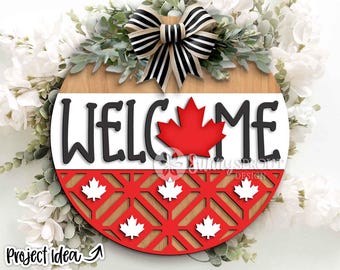 May include: A round wooden welcome sign with a Canadian theme. The sign features the word "WELCOME" in black letters, a red maple leaf, and a red and white patterned bottom. A black and white striped bow tops the sign.