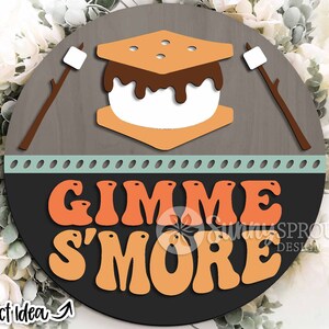 K&ouml;nnte beinhalten: Ein rundes Holzschild mit braunem Hintergrund und hellblauem Rand. Das Schild zeigt eine Grafik eines S'mores mit Schokoladen&uuml;berzug und den Text "Gimme S'more" in orangefarbenen und gelben Buchstaben.