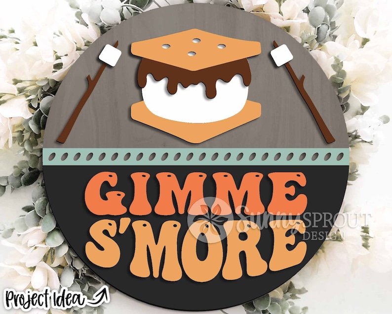K&ouml;nnte beinhalten: Ein rundes Holzschild mit braunem Hintergrund und hellblauem Rand. Das Schild zeigt eine Grafik eines S'mores mit Schokoladen&uuml;berzug und den Text "Gimme S'more" in orangefarbenen und gelben Buchstaben.