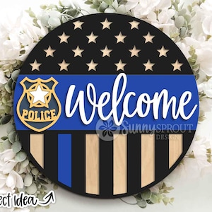 May include: Circular wooden welcome sign. The top section is black with gold stars. A blue stripe across the middle has the word "Welcome" in white. A police badge is on the left. The bottom has black and wood stripes.