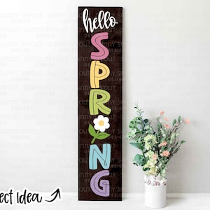 Peut inclure: Une pancarte en bois avec le mot "hello" et le mot "spring" écrits verticalement en lettres colorées. Une petite fleur avec des feuilles vertes se trouve entre les lettres "R" et "I".