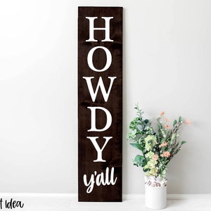 以下が含まれることがあります： 白い文字で「HOWDY y'all」と書かれた濃い茶色の木製看板。看板は白い壁に立てかけられ、その前に花瓶が置かれている。