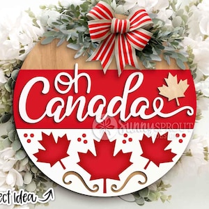 Könnte beinhalten: Runder Holzschild mit dem Schriftzug "Oh Canada" in weißer Schrift, roten und weißen Ahornblatt-Akzenten und einer rot-weiß gestreiften Schleife. Das Schild ist mit Grün und weißen Blumen verziert.