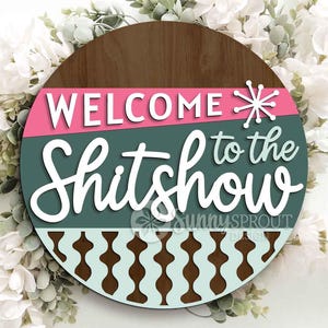 May include: Round wooden sign with the text "WELCOME to the Shitshow" in white script. The sign has a pink and teal color scheme with a brown wood grain top and a decorative pattern at the bottom.