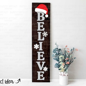 Puede incluir: Un letrero de madera con la palabra "BELIEVE" en letras blancas, con un gorro de Papá Noel rojo en la parte superior y copos de nieve por todas partes. El letrero está en una pared blanca con un jarrón de flores delante.