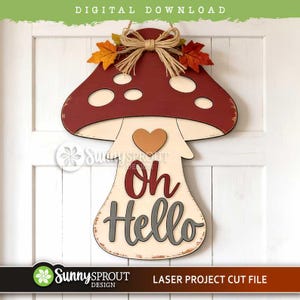 May include: A mushroom-shaped wooden wall hanging featuring a dark red cap with cream-colored spots, a cream stem, and a brown heart. The piece is topped with a jute twine bow and two fabric autumn leaves. The text Oh Hello is printed in red and gray.