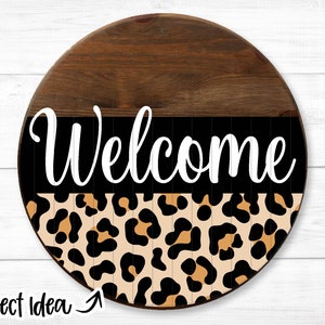 Puede incluir: Un letrero redondo de madera con un mensaje "Bienvenido" en blanco y negro y un patrón de estampado de leopardo.