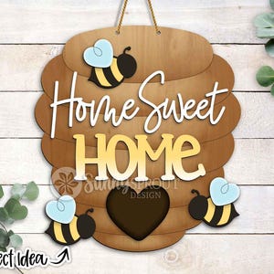 Puede incluir: Cartel de madera con diseño de colmena, con las palabras "Home Sweet Home" en blanco y amarillo. Incluye detalles de abejas y corazones. El cartel está suspendido por un cordón de hilo natural. Las palabras "Project Idea" están en la esquina inferior izquierda.