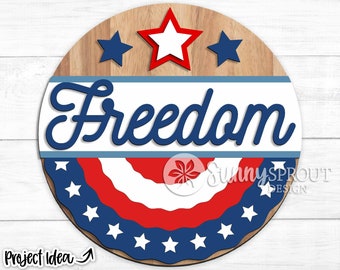 May include: A round wooden sign with a red, white, and blue American flag design. The sign says "Freedom" in blue cursive lettering. There are three stars at the top of the sign, two blue and one red.