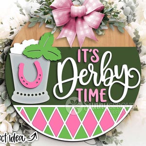 May include: A round wooden sign with a green background and a pink and green diamond pattern. The sign says "It's Derby Time" in white lettering and features a mint julep cup with a pink horseshoe.