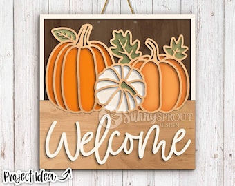 May include: Wooden welcome sign featuring layered pumpkins in orange and white, with green leaves. The word "Welcome" is in a cursive font. The sign is framed and has a neutral color scheme, perfect for fall decor.