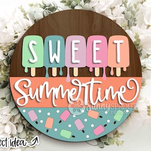May include: A round wooden sign with the words "SWEET Summertime" in white script. The word "SWEET" is written with popsicle-shaped letters in shades of green, blue, pink, and orange. The sign has a brown wood grain background and a decorative border with popsicle and dot patterns.