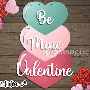 May include: Three layered paper hearts in pink, light pink, and teal with the words "Be Mine Valentine" written in white on them. The hearts are on a wood background with glitter hearts scattered around them.