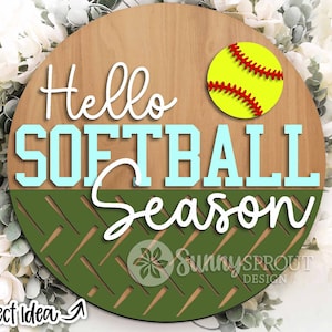 Pode incluir: Um sinal redondo de madeira com o texto "Hello Softball Season" em letras brancas e azuis. Uma bola de softball amarela com costuras vermelhas está no canto superior direito. O fundo é um padrão de chevron verde e branco.
