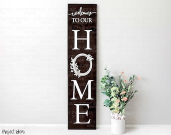 May include: A brown wooden sign with white lettering that reads "Welcome to our Home". The letters are in a vertical stack with a wreath design around the "O".