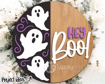 May include: A round wooden sign with a black and white ghost design on one half and the text "Hey Boo!" in white and purple on the other half.