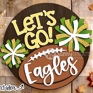 Könnte beinhalten: Ein rundes, dunkelbraunes Holzschild mit dem gelben Schriftzug "LET'S GO!" über einer braunen Football-Grafik. Das Schild enthält zwei grüne, weiße und gold glitzernde Pompon-Ausschnitte sowie den weißen Schriftzug "Eagles" auf dem Football.