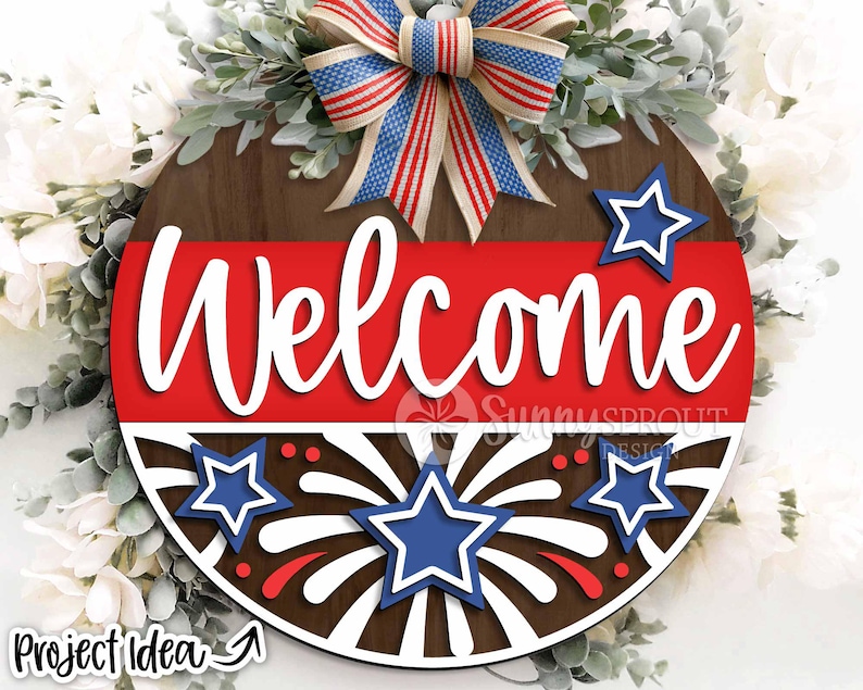 May include: A round wooden sign with a red, white, and blue patriotic theme. The sign says "Welcome" in white letters and features a firework design with stars.