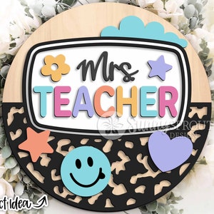 Peut inclure: Une pancarte ronde en bois avec un motif léopard noir et blanc. La pancarte présente une découpe colorée d'un écran de télévision avec le texte "Mrs. TEACHER" dans une police arc-en-ciel. La pancarte comprend également une fleur, une étoile, un cœur et un visage souriant.