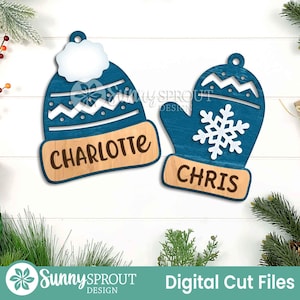 Pode incluir: Dois enfeites de Natal de madeira: um gorro de inverno azul com o nome "Charlotte" e uma luva azul com um floco de neve e o nome "Chris". Ambos os enfeites têm uma base castanha clara. São arquivos de corte digital.