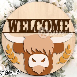 Peut inclure: Une pancarte ronde en bois avec une t&ecirc;te de vache &eacute;cossaise brune et le mot "WELCOME" en lettres brunes. La vache a un visage blanc et des cornes brunes. La pancarte est entour&eacute;e d'une couronne de tiges de bl&eacute;.