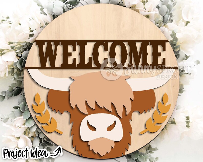 Peut inclure: Une pancarte ronde en bois avec une t&ecirc;te de vache &eacute;cossaise brune et le mot "WELCOME" en lettres brunes. La vache a un visage blanc et des cornes brunes. La pancarte est entour&eacute;e d'une couronne de tiges de bl&eacute;.