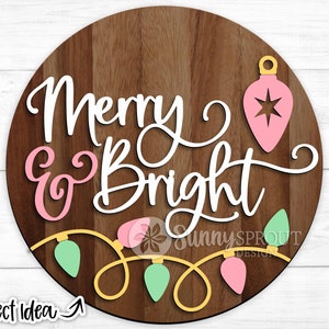 May include: A wooden round sign with the text "Merry & Bright" in white script with pink accents. The sign is decorated with a pink ornament and a string of lights in pink and green.