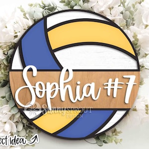 Peut inclure: Panneau rond en bois sur le thème du volley-ball avec le nom "Sophia" et le numéro "7" en écriture blanche. Le panneau présente des sections bleues, jaunes et blanches, avec un centre en grain de bois. Le texte "SunnySprout Design" est également visible.