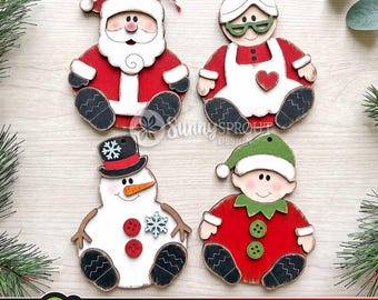 May include: Four wooden Christmas ornaments featuring Santa Claus, Mrs. Claus, a snowman, and an elf. Each character is painted in festive colors with black boots and white accents. Santa has a red and white string bow, while the snowman wears a top hat.