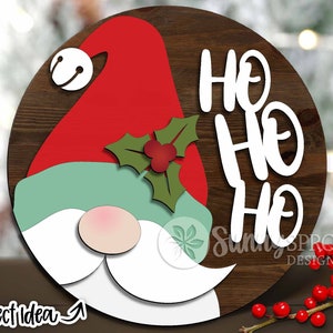 Może przedstawiać: Okrągły drewniany znak z twarzą gnoma i napisem "Ho Ho Ho" w kolorze białym. Gnom ma czerwony kapelusz, zieloną brodę i białą brodę.