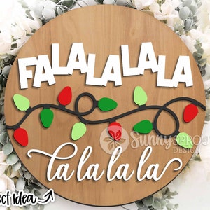 以下が含まれることがあります： 白い筆記体で「Fa La La La La la la la」と書かれた丸い木製の看板。看板は赤、緑、白のライトのストリングで飾られています。