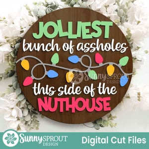 May include: A circular dark wood sign featuring the text JOLLIEST bunch of assholes this side of the NUTHOUSE in layered green, white, and pink lettering. A string of colorful yellow, blue, pink, and green Christmas light bulbs is centered.