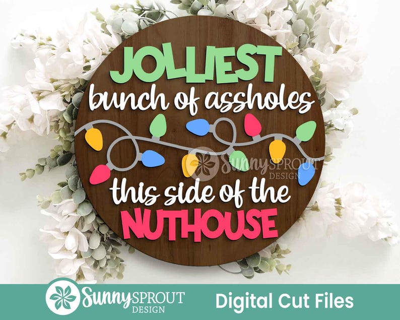 May include: A circular dark wood sign featuring the text JOLLIEST bunch of assholes this side of the NUTHOUSE in layered green, white, and pink lettering. A string of colorful yellow, blue, pink, and green Christmas light bulbs is centered.