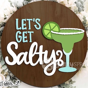 May include: A round wooden sign with the text "Let's Get Salty" in blue and white lettering. A green margarita glass with a lime wedge is pictured next to the text.