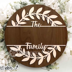 May include: A round wooden sign with the words "The Family" cut out. The sign is brown with a light-colored leaf design around the top and bottom. The words are in a cursive font.