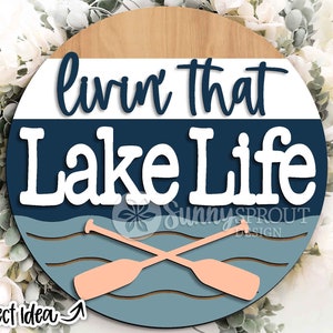 Op de afbeelding: Een rond houten bord met de tekst "Livin' that Lake Life" in witte letters op een blauwe achtergrond. Het bord heeft een houtnerfstructuur en een lichtbruine rand. Twee gekruiste roeispanen zijn afgebeeld in de onderste helft van het bord, met een blauw en wit golfpatroon erachter.