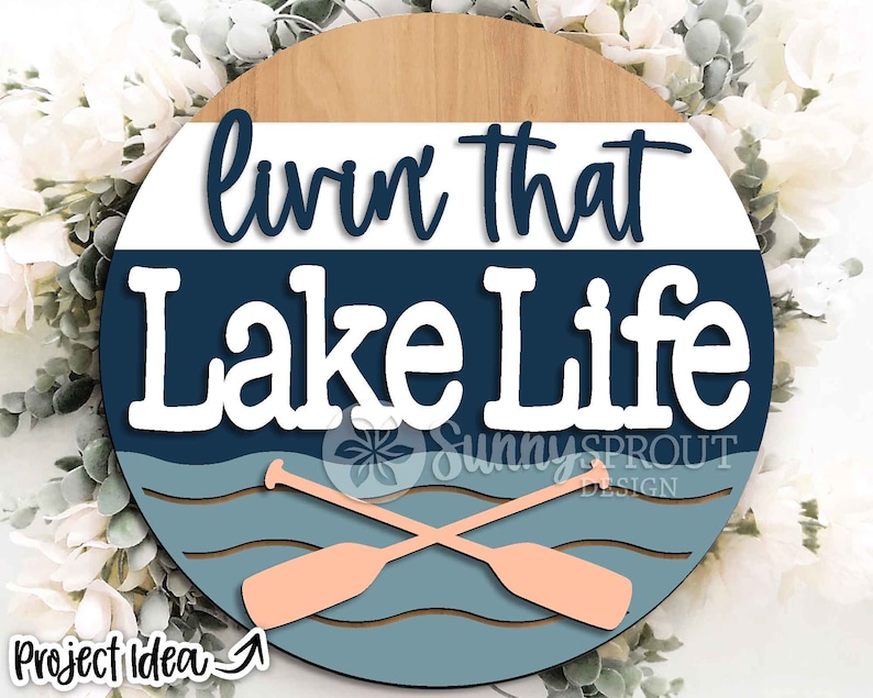 Op de afbeelding: Een rond houten bord met de tekst "Livin' that Lake Life" in witte letters op een blauwe achtergrond. Het bord heeft een houtnerfstructuur en een lichtbruine rand. Twee gekruiste roeispanen zijn afgebeeld in de onderste helft van het bord, met een blauw en wit golfpatroon erachter.