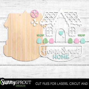 May include: Wooden house-shaped cutout with a decorative white house overlay. The overlay features windows, a heart, and the words "Home Sweet Home." Accents include pink, green, and blue elements. The design is for laser cutting and crafting.