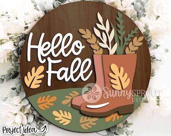 May include: A round wooden sign with the text "Hello Fall" in white lettering. The sign is decorated with fall-themed elements, including a pair of brown rain boots with a leafy design, and orange and yellow leaves.