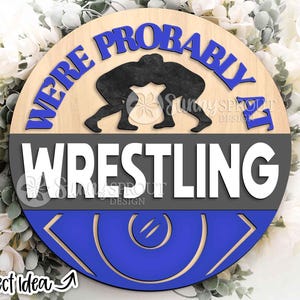Puede incluir: Un letrero redondo de madera con el texto "WE'RE PROBABLY IT WRESTLING" en azul y blanco. El letrero presenta una silueta negra de dos luchadores. La sección inferior es azul con un diseño decorativo. "Project Idea" está escrito en la parte inferior.
