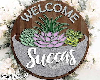 May include: A round wooden sign with the words "Welcome Succas" in white lettering. The sign has a gray background with a textured look and features a green and pink succulent plant in a gray pot.