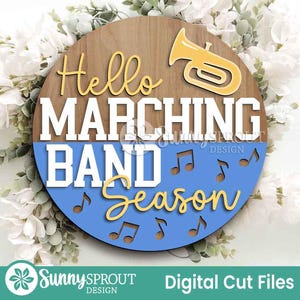 May include: A round wooden sign with the words "Hello MARCHING BAND Season" in white and yellow lettering. A yellow trumpet graphic is at the top. The bottom half is blue with musical notes. The sign is surrounded by white flowers and greenery.