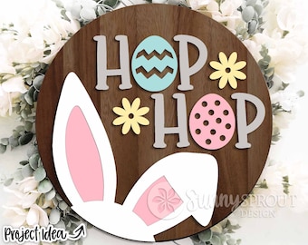 Peut inclure: Une pancarte ronde en bois avec les mots "Hop Hop" en lettres grises, décorée d'œufs de Pâques roses et bleus et de fleurs jaunes. La pancarte présente également un lapin blanc avec des oreilles roses.